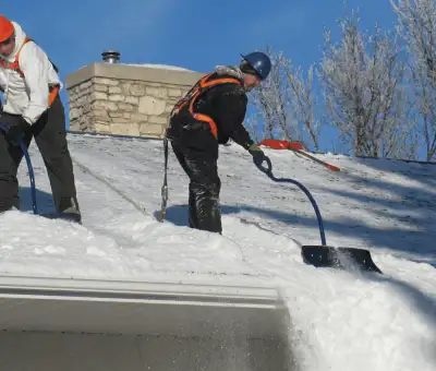 Professional technicians performing roof snow removal with safety harnesses and shovels in Spokane, WA